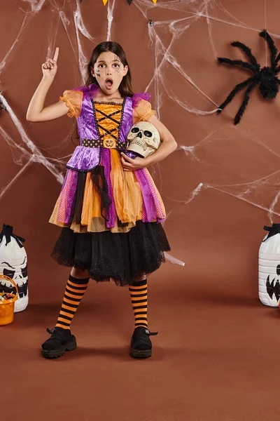 Chica impactada en vestido de Halloween de pie con el cráneo y apuntando hacia arriba con el dedo sobre fondo marrón - foto de stock
