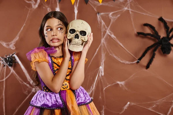 Menina em traje de Halloween em pé com crânio e sorrir no fundo marrom, estação assustadora — Fotografia de Stock