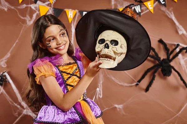 Glückliches Mädchen in Halloween-Kleidung mit Hexenhut auf Totenkopf auf braunem Hintergrund, gruselige Jahreszeit — Stockfoto