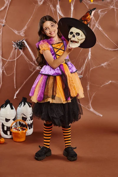Chica feliz en disfraz de Halloween con sombrero de bruja en el cráneo sobre fondo marrón, temporada espeluznante - foto de stock
