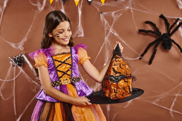Menina feliz em Halloween bruxa fantasia segurando chapéu pontudo no fundo marrom, estação assustadora — Fotografia de Stock