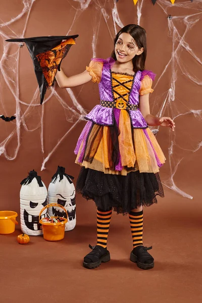 Cheerful child in Halloween witch costume holding pointed hat upside down on brown background — Stock Photo