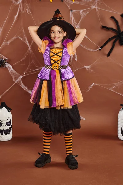 Fille heureuse en chapeau de sorcière et costume d'Halloween debout à côté d'un décor effrayant bricolage sur fond brun — Photo de stock