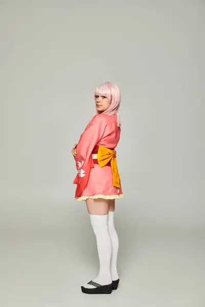 Joven mujer de estilo anime en peluca rubia y kimono rosa con lazo amarillo sobre gris, concepto cosplay - foto de stock