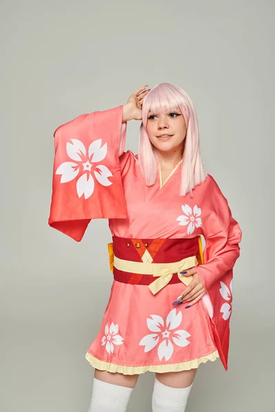Joven mujer sonriente en peluca rubia y kimono rosa con estampado floral en gris, estilo anime - foto de stock