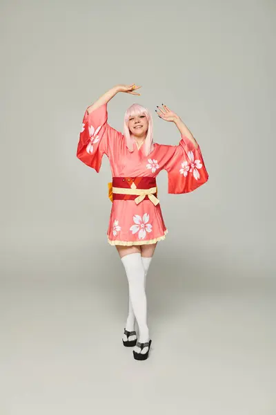 Mujer rubia cosplay en kimono colorido y calcetines blancos de rodilla posando con las manos levantadas en gris - foto de stock