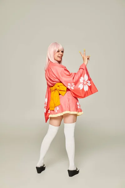 Jovem mulher em peruca loira e quimono rosa com meias de joelho branco mostrando gesto de vitória em cinza — Fotografia de Stock