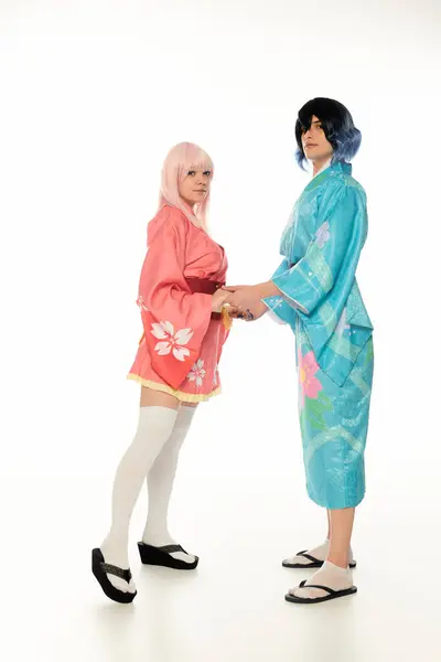 Молодая пара в красочных кимоно и париках, держась за руки и глядя на камеру на белом, в стиле аниме — стоковое фото