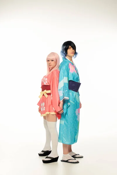 Giovani cosplayers in kimono colorati e parrucche in piedi schiena contro schiena e tenendo le mani sul bianco — Foto stock