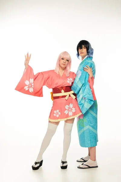 Jeune femme en kimono agitant la main près anime style homme en perruque sur blanc, asiatique sous-culture mode — Photo de stock
