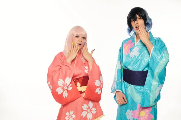 Asombrada pareja de anime en coloridos kimonos y pelucas tocando caras en blanco, tendencia cosplay - foto de stock