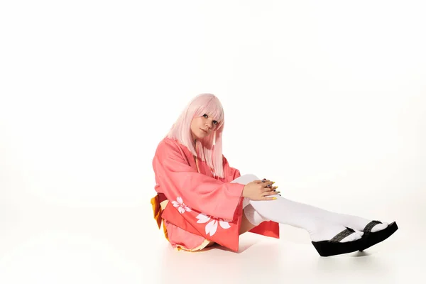 Charming woman in pink kimono and blonde wig sitting and looking at camera on white, anime style — Stock Photo