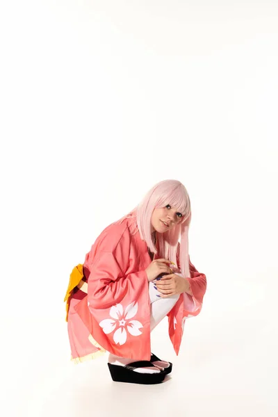 Young anime woman in pink kimono and blonde wig sitting on haunches and looking at camera on white — Stock Photo