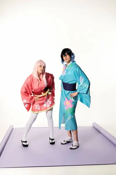 Молодые игривые косплееры в кимоно и париках с руками на бедрах на фиолетовом ковре в белой студии — стоковое фото