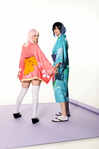 Anime style couple en kimonos tenant la main et regardant la caméra sur tapis violet en studio blanc — Photo de stock
