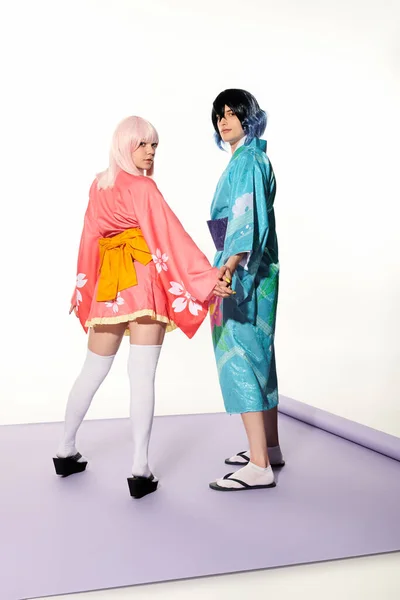 Anime style couple in kimonos holding hands and looking at camera on purple carpet in white studio — Stock Photo
