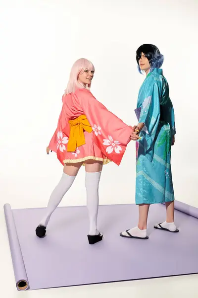 Jovem casal em quimonos e perucas de mãos dadas e sorrindo para a câmera no tapete roxo no branco — Fotografia de Stock