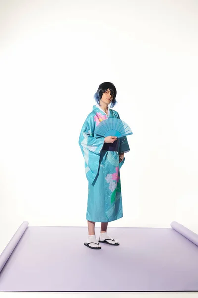 Uomo stile anime in kimono blu e parrucca tenendo ventilatore a mano e guardando la fotocamera su sfondo bianco — Foto stock