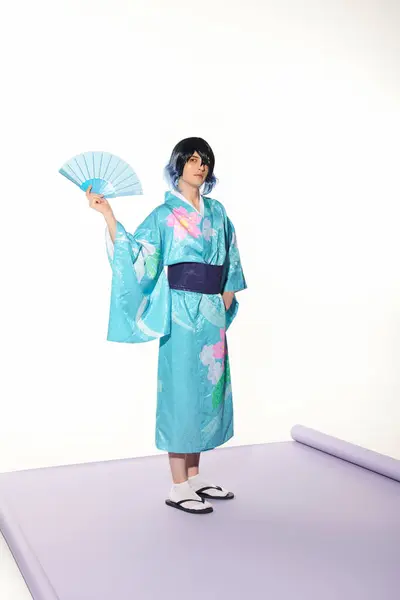 Косплей-стиль, человек в синем кимоно и парике, позирующий с веером на фиолетовом ковре и белом рюкзаке — стоковое фото