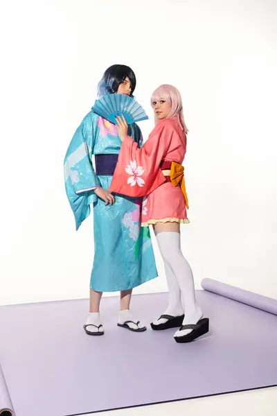 Блондинка в стиле аниме, держащая за руку вентилятор рядом с мужчиной в кимоно на фиолетовом ковре в белой студии — стоковое фото