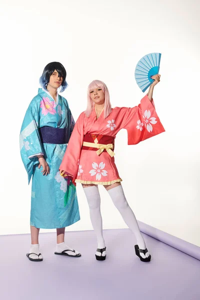 Anime style woman posing with hand fan near man in kimono and wig on purple carpet in white studio — Stock Photo