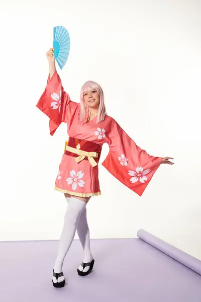 Charming anime style woman in pink kimono and wig standing with hand fan in artistic pose on white — Stock Photo