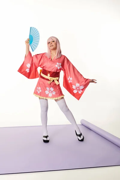 Mulher cosplay artístico em quimono rosa e peruca loira com ventilador de mão no tapete roxo no estúdio branco — Fotografia de Stock