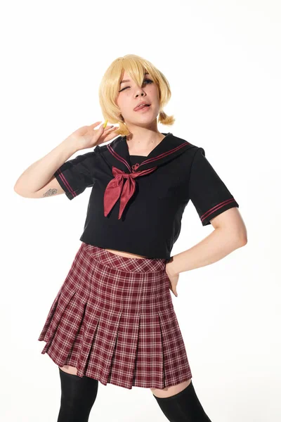 Seductora mujer estilo anime en peluca rubia amarilla y uniforme escolar posando con la mano en la cadera sobre blanco - foto de stock