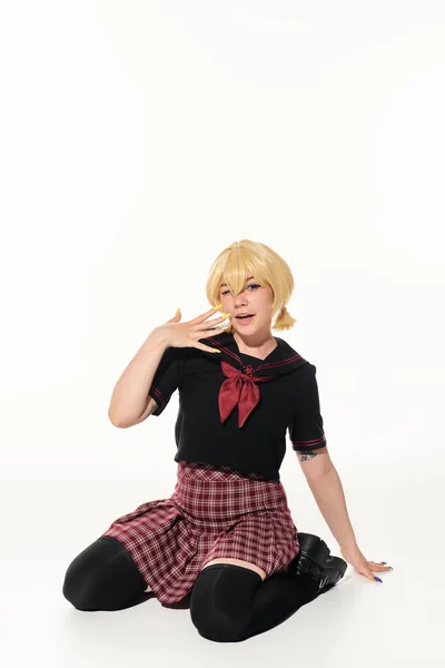 Стильная блондинка в школьной форме показывает блондинку парик, сидя на белом, аниме характер — стоковое фото