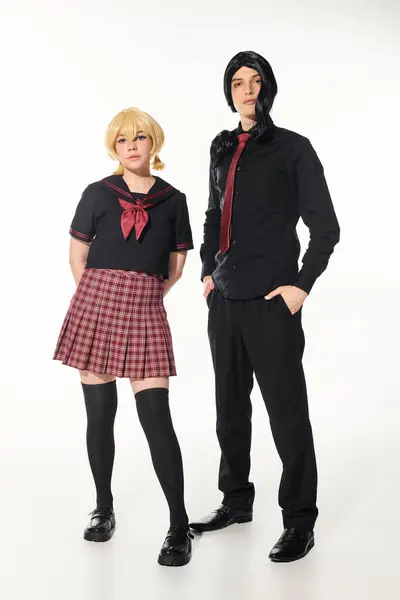 Uomo in abiti neri con le mani in tasca vicino alla donna bionda in uniforme scolastica su bianco, cosplayers — Foto stock