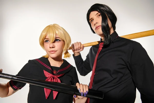 Young cosplayers in blonde and black  wigs posing in dark school uniform with baseball bats on grey — Stock Photo