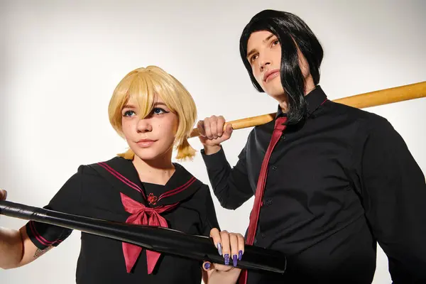 Giovani cosplayers in parrucche bionde e nere in posa in uniforme scuola scura con mazze da baseball su grigio — Foto stock