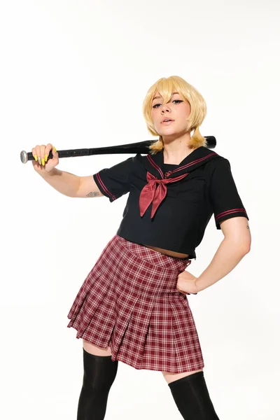 Cosplay характер, надменная женщина в школьной форме с бейсбольной битой и рукой на бедре на белом — стоковое фото