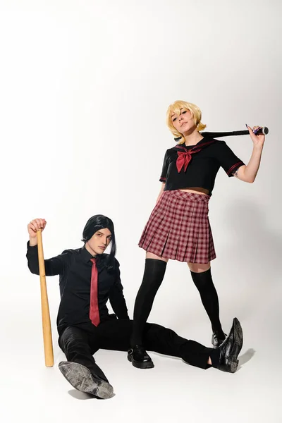 Homme en perruque noire assis avec batte de baseball près de la femme en uniforme scolaire sur blanc, tendance cosplay — Photo de stock