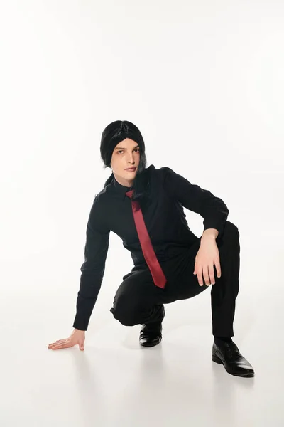Anime man in black clothes and wig with red tie sitting on haunches looking at camera on white — Stock Photo