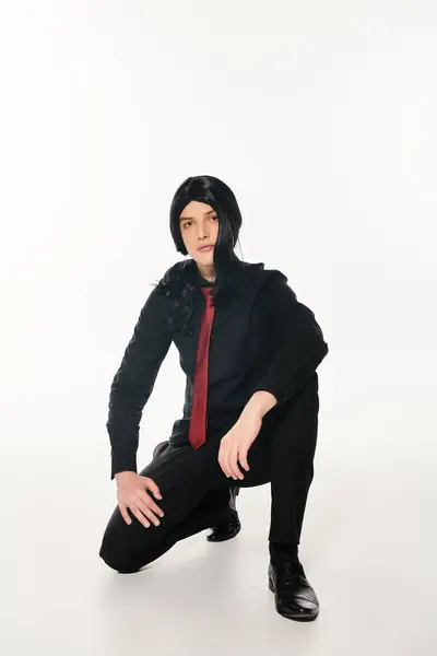 Cosplay man in black clothes and wig with red tie posing on haunches looking at camera on white — Stock Photo