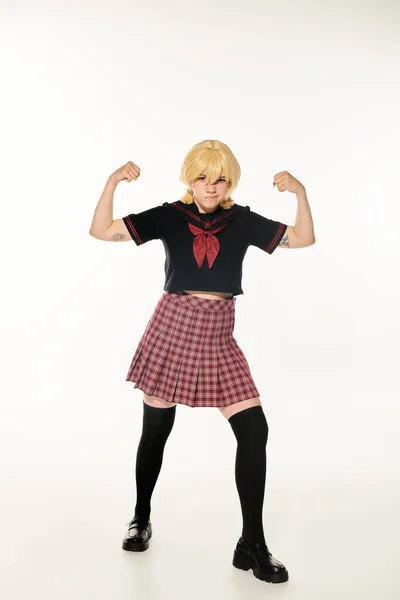 Wütend unzufriedene Frau im Anime-Stil in Schuluniform und gelber blonder Perücke, die Muskeln auf Weiß zeigt — Stockfoto
