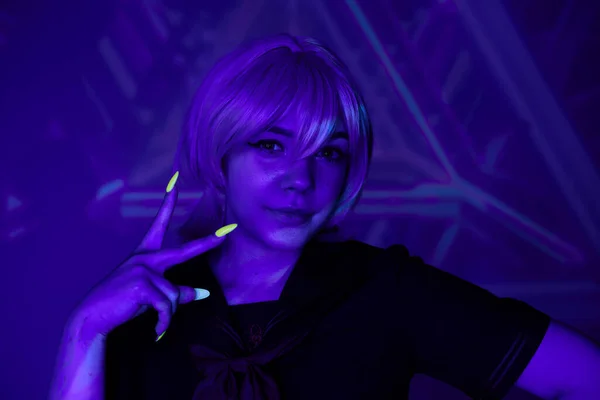 Mujer de estilo anime con peluca rubia y manicura fluorescente mostrando signo de victoria en luz de neón azul - foto de stock