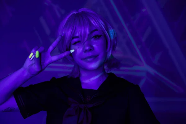 Donna anime bionda con manicure fluorescente strizzando l'occhio e mostrando segno di vittoria in luce blu al neon — Foto stock