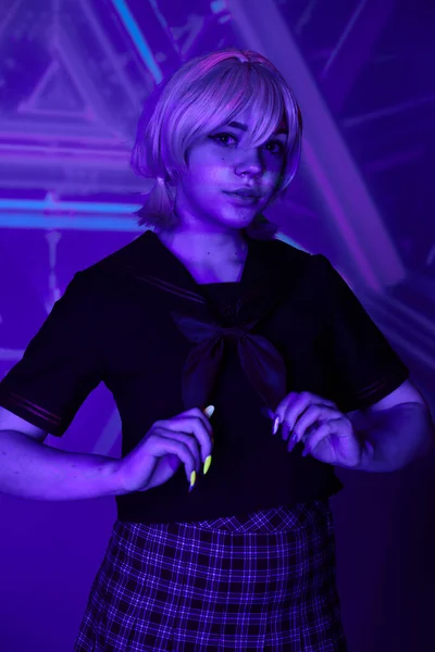 Giovani cosplay cultura, donna in parrucca bionda e uniforme scolastica guardando la fotocamera in luce blu al neon — Foto stock