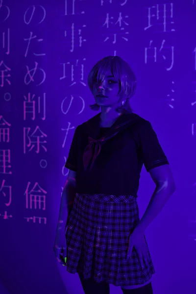 Cosplay-Frau in Schuluniform steht in blauem Neonlicht vor Hintergrund mit Hieroglyphen-Projektion — Stockfoto