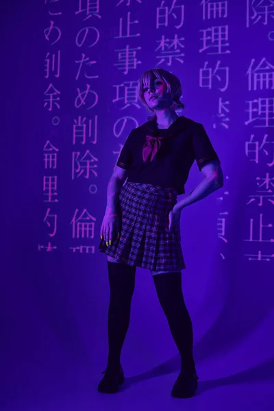 Mujer de estilo anime en uniforme escolar con mano en la cadera en luz de neón azul con proyección de jeroglíficos - foto de stock
