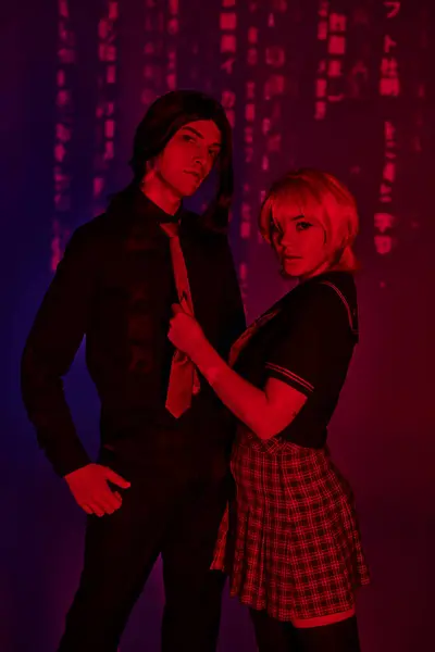 Anime couple in students  looking at camera in red neon light on abstract purple backdrop — Stock Photo