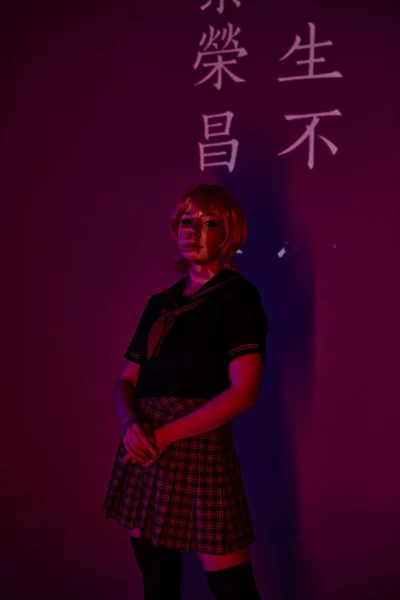 Anime-Frau in Perücke und Schuluniform in Neonlicht vor violettem Hintergrund mit Hieroglyphen-Projektion — Stockfoto