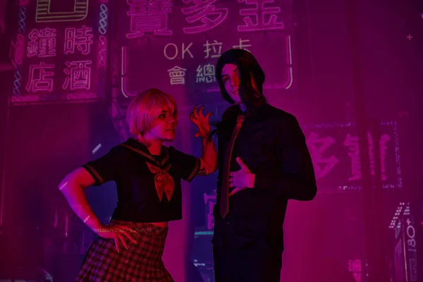 Young woman in school uniform scaring anime style boyfriend on purple neon backdrop with hieroglyphs — Stock Photo