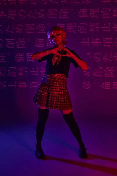 Stylish anime woman in school uniform showing heart sign on neon purple backdrop with hieroglyphs — Stock Photo