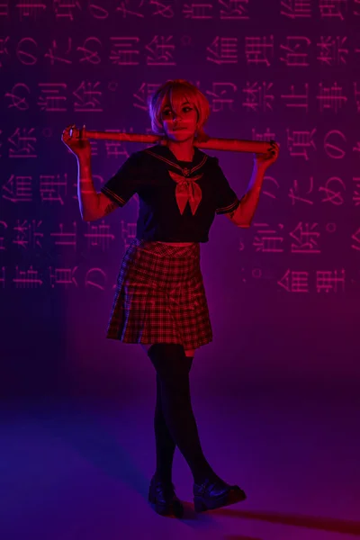 Trendy cosplay woman in school uniform with baseball bat on neon purple backdrop with hieroglyphs — Stock Photo