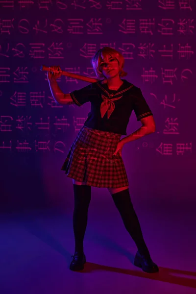 Cosplay woman in school uniform posing with baseball bat on neon purple backdrop with hieroglyphs — Stock Photo