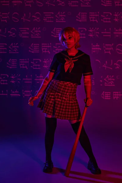 Cosplay donna in uniforme scolastica in piedi con mazza da baseball su sfondo viola neon con geroglifici — Foto stock