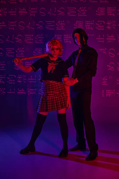 Anime woman with baseball bat near man in wig on neon purple backdrop with hieroglyphs projection — Stock Photo
